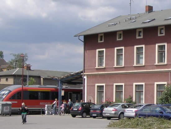 Seitenansicht des Bahnhofes in Kamenz. Eine Regionalbahn steht am Gleis. Menschen stehen davor und steigen ein bzw. aus. Auf dem Parkplatz stehen verschiedene Autos.