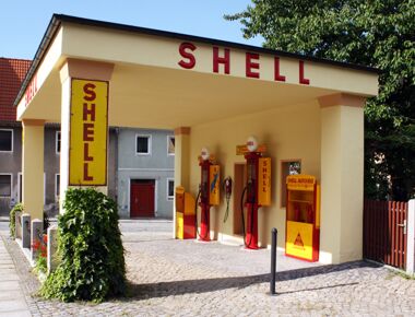 Außenansicht der historischen Schell-Tankstelle in Kamenz. Ein gelbes Gebäude mit der Aufschrift Schell und zwei rot-gelben Tankzapfsäulen im Mittelpunkt. Beispiel für das Tankstellenmuseum in Kamenz.
