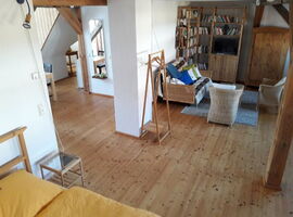 Großer offener Wohnbereich mit Holzdielen und Balken. Rechts Bücherregale, Fernseher, Schrank, Korbsofa, Sesseln und Tisch. Links Treppenaufgang und Sitzgruppe. Im Vordergrund Bett mit Beistelltisch.. 