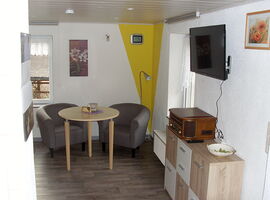 Wohnbereich in der FeWo Voigt. Zwei Sessel mit einem runden Tisch und einer Lampe stehen in einer Raumecke zwischen zwei Fenstern. Rechts davor eine Kommode in Holzoptik mit einem Nostalgie-Radio darauf und einem TV darüber. Blumen-Bilder an den Wänden.. 