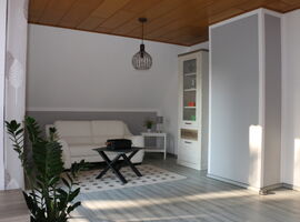 Blick in anderen Bereich des Wohnzimmers nahe Balkon. An der Wand steht eine beige-farbene Ledercouch mit einem schwarzen Couchtisch davor. Darunter ein karierter Teppich. Rechts ein Beistelltisch mit Blumenschmuck und Lampe. Daneben ein Schrank mit Deko.. 