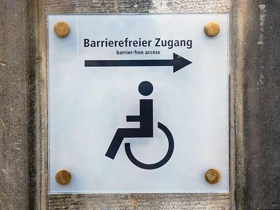 Ein Schild an einer Wand mit einem Strichmännchen-Symbol in einem Rollstuhl und der Aufschrift: "Barrierefreier Zugang, barrier-free access". Symbolbild für Barrierefreiheit und barrierefreie Einrichtungen.. 