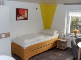 Ein Einzelbett aus Holz mit einem Nachttisch und einer Lampe daneben. Das Bett ist möglicherweise erweiterbar für eine weitere Person.