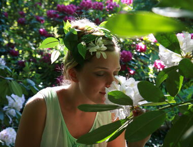 Eine Gartenfee (Model) im Kleid, mit Blumen im Haar riecht an den Rhododendronblüten auf dem Kamezer Hutberg. Alles ist von Blättern und weißen und rosafarbenen Blüten umgeben. Die Sonne scheint durch die Blätter. Symbolbild für Natur in Kamenz genießen.. 