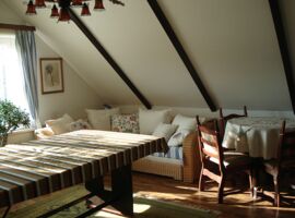 Einblick Wohnraum mit einem großen Tisch links mit Decke darauf. Im Hintergrund ein Fenster mit Sonneneinstrahlung und Zimmerpflanze. Rechts ein Korbsofa mit zahlreichen Kissen. Rechts danben ein runder Tisch mit Deckchen und Holzstühlen.