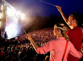 Ein riesiges Publikum vor der Hutberg-Freilichtbühne in Kamenz während eines Konzerts. Im Vordergrund zwei Konzertbesucher mit Dirigentenstäben. Beispielbild für Events in Kamenz.. 