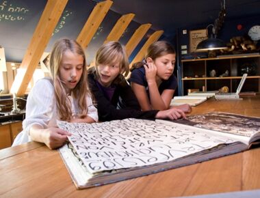 Drei Kinder sehen sich gemeinsam ein großes Buch mit Schriftzeichen im Museum der Westlausitz an. Beispielbild für das Stöbern in der Themenwelt Idee im Elementarium. Im Hintergrund sind zahlreiche Exponate ausgestellt.