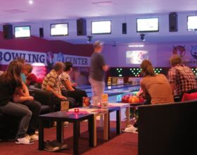 Innenansicht des Bowling-Center Kamenz. Verschiedene Personen unterschiedlichen Alters spielen zusammen Bowling. Aktueller Spieler bereitet sich auf seinen Wurf vor, während die anderen an Tischen zuschauen und warten. Beispiel für Freizeiteinrichtungen.