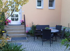 Sitzgruppe auf einer Terrasse im Gartenbereich. Um einen runden Tisch stehen vier Stühle und eine Sitzbank. Links ist eine Eingangstür zum Haus mit Treppenaufgang und Blumenschmuck.. 