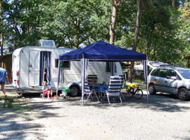 Beispiel für ein Wohnmobil auf dem Campingplatz Deutschbaselitz. Ein Wohnmobil mit offener Tür, an der eine Frau sitzt. Davor stehen ein Pavillon-Zelt und ein Campingtisch mit vier Stühlen. Rechts stehen ein Fahhrad und ein Auto. Im Hintergrund Bäume.