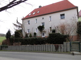 Hausansicht der Ferienwohnung Frenzel. Ein Vier-Parteien-Haus mit Treppenzugang und Hecke davor. Im Hintergrund eine Wiese und Bäume. Im Vordergrund eine Straße mit Gehweg und einer Katze.. 
