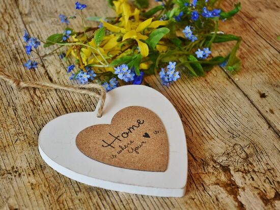 Ein Holz- / Korkherz mit der Aufschrift "Home is where your heart is" übersetzt "Zuhause ist, wo das Herz ist". Es liegt auf einem Holztisch mit Wildblumen. Symbolbild für Wohnen und Heim.Holztisch. 