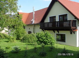 Alternative Außenansicht des Hauses mit einem großen Balkon. Davor eine grüne Wiese mit zahlreichen Sträuchern und kleinen Nadelbäumen.. 