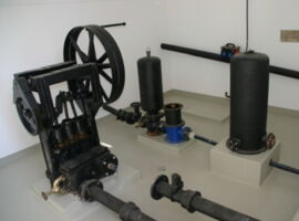 Ansicht der historischen Wasserpumpanlage Hydraulischer Widder. Verschiedene schwarze Anlagenteile, Rohre und Ventile sind miteinander verbunden in einem grau-weißen Raum. Beispielansicht für das Denkmal Hydraulischer Widder in Kamenz.