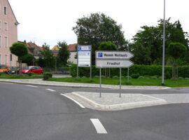 Ansicht des Kreisverkehres Böhnischplatz in Kamenz mit Zufahrt zum Stellplatz. Ausfahrt nach rechts mit der Beschilderung Parkplatz Museum Westlasuitz und Friedhof. Im Hintergrund Gebäude, Autos, Grünflächen und weitere Schilder.