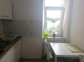 Küche mit Küchenzeilte und Spüle links. Rechts ein Esstisch mit zwei Stühlen. Im Hintergrund ein Fenster mit einer Orchidee und einer Gießkanne.. 