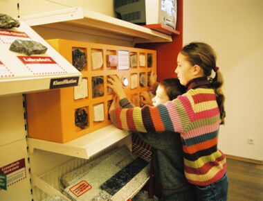 Zwei Kinder sehen sich gemeinsam verschiedene an einer Wand ausgestellte Steine an und fühlen die Formen mit ihren Händen. Beispiel für die Themenwelt Steine.