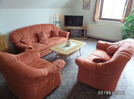 Wohnzimmer mit Teppich. Im Mittelpunkt eine Sitzgruppe mit zwei Polstersofas, Kissen und einem Sessel. Daziwschen ein Holzcouchtisch mit Milchglas. Links eine Anrichte. Im Hintergrund Eckschrank mit TV, Kerzen, eine Pflanze und ein Fenster.. 