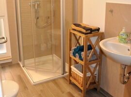Badezimmer mit Bodenbelag in heller Holzdielenoptik. Auf der linken Seite Toilette und Fenster. Rechts nahezu ebenerdige Dusche, Holzregal mit Handtüchern und Korb und ein Waschbecken.. 