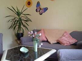Beispiel Wohnzimmer mit einem Zweier-Sofa im Hintergrund. Rechts und links davon ist jeweils ein Sessel. In der Mitte der Sitzgruppe steht ein Fliesen-Ecktisch mit Dekoschale und Blumenvase darauf. An der Wand Dekoration in Blumen- und Schmetterlingform.. 