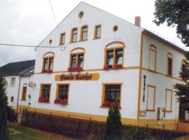 Außenansicht von Fissels Gasthof. Ein großes Gasthaus mit Blumenschmuck an den Fenster und der Aufschrift Fissels Gasthof als großes Logo auf der Wand. Im Vordergrund ein Gartenbereich mit Hecke und Sträuchern.