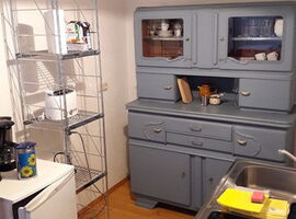 Küchenbereich mit einem Kühlschrank, Kaffeemaschine und einem Metall-Regall und Toaster links. Rechts im Hintergrund ein restaurierter Vintage-Küchenschrank. Rechts im Vordergrund eine kleine Pantry-Küche mit Spüle und zwei Herdplatten.. 