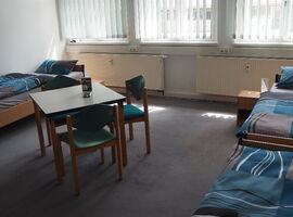 Zimmerbeispiel des Hostel jo-si mit drei Einzelbetten und Nachttischen mit Lampe an den Wänden. In der Mitte ein Tisch mit drei Stühlen und Aufsteller. Im Hintergrund drei große Fenster mit Vorhängen.. 