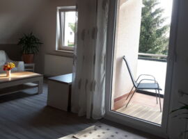 Zweite Wohnzimmer Ansicht mit Blick auf den Balkonbereich. Rechts Balkontür mit Vorhängen und Stuhl auf dem Balkon. Links eine weiße Ledercouch mit Couchtisch und Blumen darauf. In der Ecke eine Palmenpflanze im Topf.