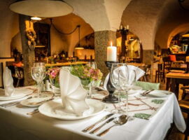 Ein festlich eingedeckter Tisch im historischen Ratskeller Restaurant des AKZENT Hotel Goldner Hirsch. Auf dem Tisch stehen vier Gedecke mit Tellern, Gläsern, Servietten, Besteck. In der Mitte Blumenschmuck. Im Hintergrund weitere Tische.