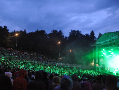 Seitenaufnahme Konzert auf der Hutbergbühne Kamenz. Eine Band spielt rechts auf der beleuchteten Bühne während der Abenddämmerung. Davor steht ein Publikum mit unzähligen Besuchern. Beispiel für Veranstaltung auf dem Hutberg.