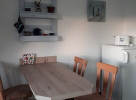 Esstisch im Küchenbereich der FeWo Am Heidelberg. Ein Esstisch in Holoptik, vier Holzstühöe, ein Wandregal mit Teller, Deko und Topflappen. Rechts ein mittelgroßer Kühlschrank mit Toaster darauf.. 