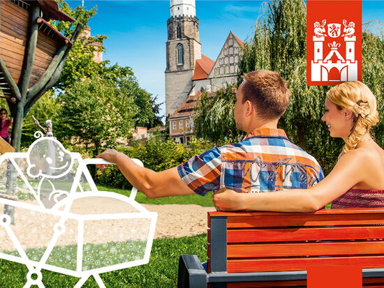 Imagebild der Kampagne Neue Stadt. Neues Glück. Ein junges Elternpaar sitzt in einem Park auf einer Bank und schaukelt einen virtuell eingezeichneten Kinderwagen links mit einem Baby darin. Im Hintergrund die Kamenzer Altstadt.. 