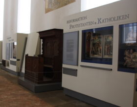 Innenansicht des Ausstellungsraumes im Sakralmuseum / Klosterkirche St. Annen. Ausstellungsteil Reformation Protestanten und Katholiken. Verschiedene Gemälde und Skulpturen in Glasvitrinen sowie Teile der ehemaligen Kirch-Innenausstattung daneben.