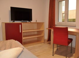 Ein helles Beispielzimmer mit einem Bett. Mittig im Hintergrund ein Schrank mit einem TV und Duftstäbchen darauf. Rechts ein kleiner Schreibtisch mit rotem Stuhl vor einem Fenster mit Vorhängen.. 