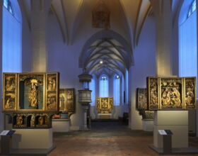 Innenansicht der Klosterkirche St. Annen / Sakralmuseum. Ausstellungsraum mit jeweils 2 beleuchteten Schnitzaltären am Rand und einem Altar im Hintergrund in der Mitte. Die Altäre veranschaulichen unter anderem Maria, Jesus und Franz von Assisi.