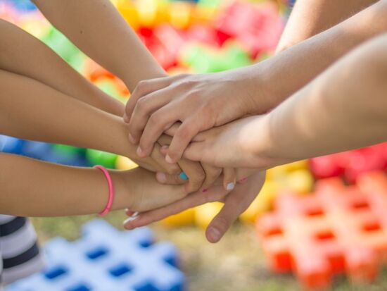 Sieben Hände sind übereinander gelegt als Zeichen des Zusammenhaltes, ähnlich wie bei dem Motto: Einer für alle - alle für einen. Kinderhände vor einem bunten Hintergrund. Symbolbild für Zusammenhalt.