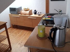 Küchenbereich unter Dachschräge mit Fenster. Links ein Regal. Im Hintergrund ein Küchenschrank mit Mini-Ofen, Brotkasten und Utensilien. Rechts eine Pantry-Küche mit Spüle und Herd sowie Kühöschrank, Kaffeemaschine und Wasserkocher. Wandbild Stadtansicht.. 