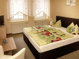 Schlafzimmer Beispiel in der Pension Kiautschau. Rechts ein Doppelbett an der Wand, bezogen mit Schmetterlingsbettwäsche. Darüber ein Bild an der Wand. Links eine Kommode mit TV sowie ein Tisch mit Teelicht und Sessel. Im Hintergrund zwei Fenster.. 