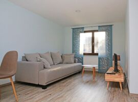 Wohnbereich mit hellem Laminat und einem Schalenstuhl links. Dahinter ein sandfarbenes Sofa mit drei Sitzflächen. Davor ein kleiner Couchtisch vor dem Fenster mit blauen Design-Vorängen. Rechts an der Wand ein TV-Reck mit TV und kleiner Lampe.