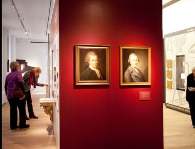 Innenansicht des Lessing-Museums mit zwei Gemälden im Mittelpunkt, auf denen der Aufklärer und Schriftsteller Lessing zu sehen ist. Drei Besucherinnen sehen sich die weiteren Ausstellungsstücke rechts und links im Raum an.. 