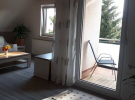 Zweite Wohnzimmer Ansicht mit Blick auf den Balkonbereich. Rechts Balkontür mit Vorhängen und Stuhl auf dem Balkon. Links eine weiße Ledercouch mit Couchtisch und Blumen darauf. In der Ecke eine Palmenpflanze im Topf.. 
