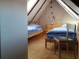 Zimmer unter Dachschrägen mit zwei Holz-Einzelbetten unter einem Dachfenster. Im Hintergrund eine gemeinsame Kommode zwischen den Betten. Im Vordergrund ein Holzstuhl. An der Wand eine Art Fee als Wandtattoo.. 