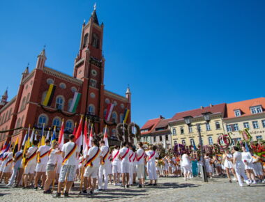 Umzug der Schüler und Lehrer zum traditionellen Forstfest. Sie tragen weiße Kleidung, Blumenschmuck, Fahnen und Schärpen und versammeln sich zum gemeinsamen Singen und Weiterzug auf dem Marktplatz vor dem Rathaus.