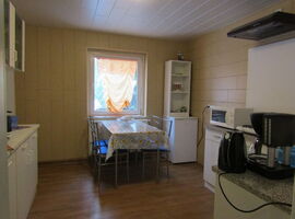 Links Küchenschränke. In der Mittel ein Metallküchentisch mit Tischdecke und Schale vor einem Fenster mit Gardine. Rechts daneben ein Schrank mit Kühlschrank. Rechts vorne eine Küchenzeile mit Mikrowelle, Herdplatten, Wasserkocher und Kaffeemaschine.. 