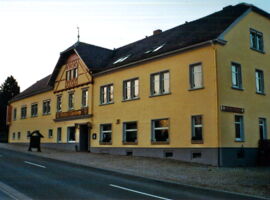 Außenansicht des Gasthof Thonberg von der Straße aus Richtung Eingangstür. Ein großes, breitgezogenes Gebäude mit drei Etagen in gelb mit einem beleuchteten Schild über der Eingangstür in der Mitte, Aufschrift Gasthof Thonberg. Angebotstafel vor dem Haus.