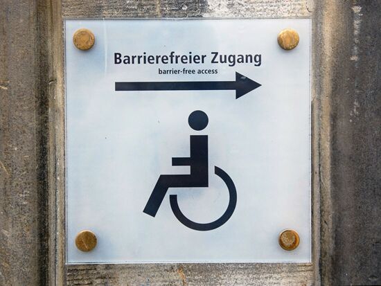 Ein Schild an einer Wand mit einem Strichmännchen-Symbol in einem Rollstuhl und der Aufschrift: "Barrierefreier Zugang, barrier-free access". Symbolbild für Barrierefreiheit und barrierefreie Einrichtungen.
