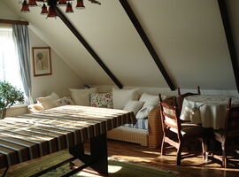 Einblick Wohnraum mit einem großen Tisch links mit Decke darauf. Im Hintergrund ein Fenster mit Sonneneinstrahlung und Zimmerpflanze. Rechts ein Korbsofa mit zahlreichen Kissen. Rechts danben ein runder Tisch mit Deckchen und Holzstühlen.. 