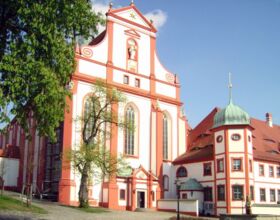 Ansicht des Kirchgebäudes im Kloster St. Marienstern in Panschwitz-Kuckau in der Nähe von Kamenz. Ein großes, weißes Gebäude mit orangefarbenen und goldenen Verzierungen. Rechts ein Turm. Links davor Bäume im Klosterhof. Beispiel benachbarte Ausflugsziele
