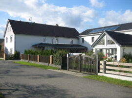 Gesamtansicht des modernen Hauses bei Sonnenschein. Grundstück ist durch Holzzäune und Doppelpforte eingegrenzt. Dazischen Gartenbereich mit Hecke, Rasen und Pflanzen. Haus mit einigen Glasfronten und Solarpanels auf dem Dach.