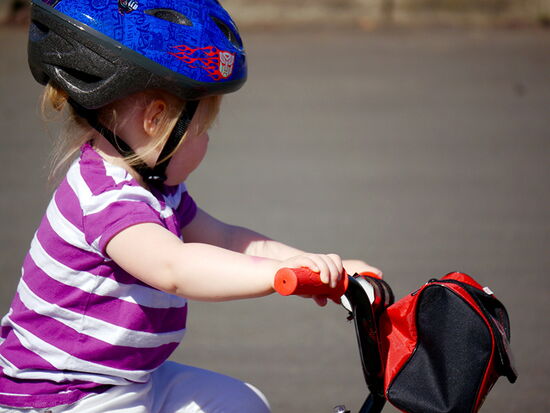 Ein Kleinkind fährt mit Helm auf einem kleinen Rad / Roller mit Lenkertasche und schaut auf die Straße. Symbolbild für Kindersicherheit.. 